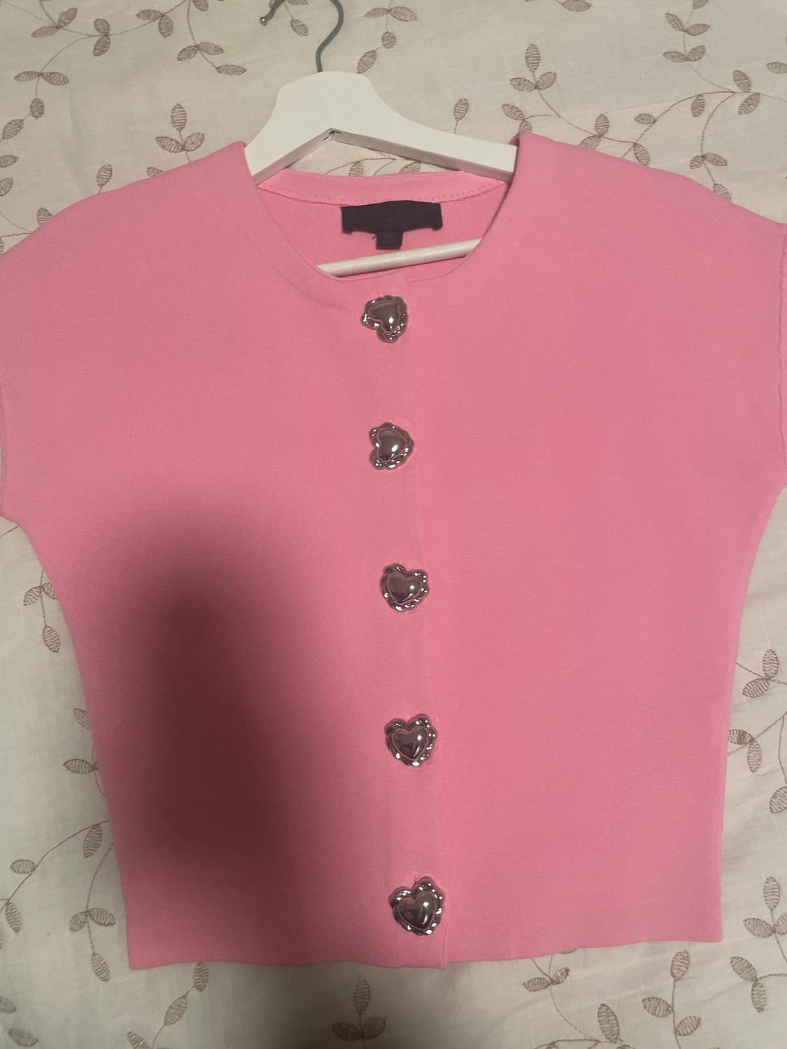 Pink jacket with silver heart-shaped buttons, hanging on a white hanger over a leaf-patterned fabric.