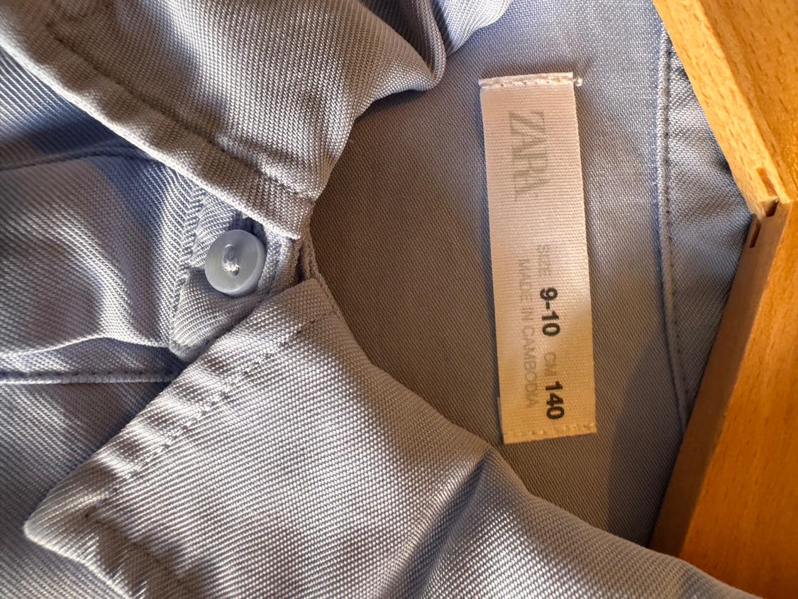 Light blue shirt with a label that reads "ZARA, 9-10, CM 140, MADE IN CAMBODIA", hanging on a wooden hanger.