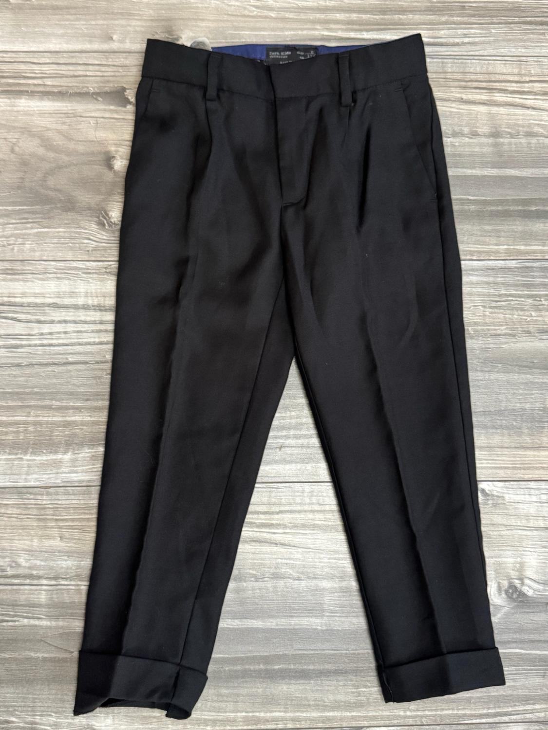Black pleated trousers with belt loops and turn-ups, photographed on a light wooden surface.