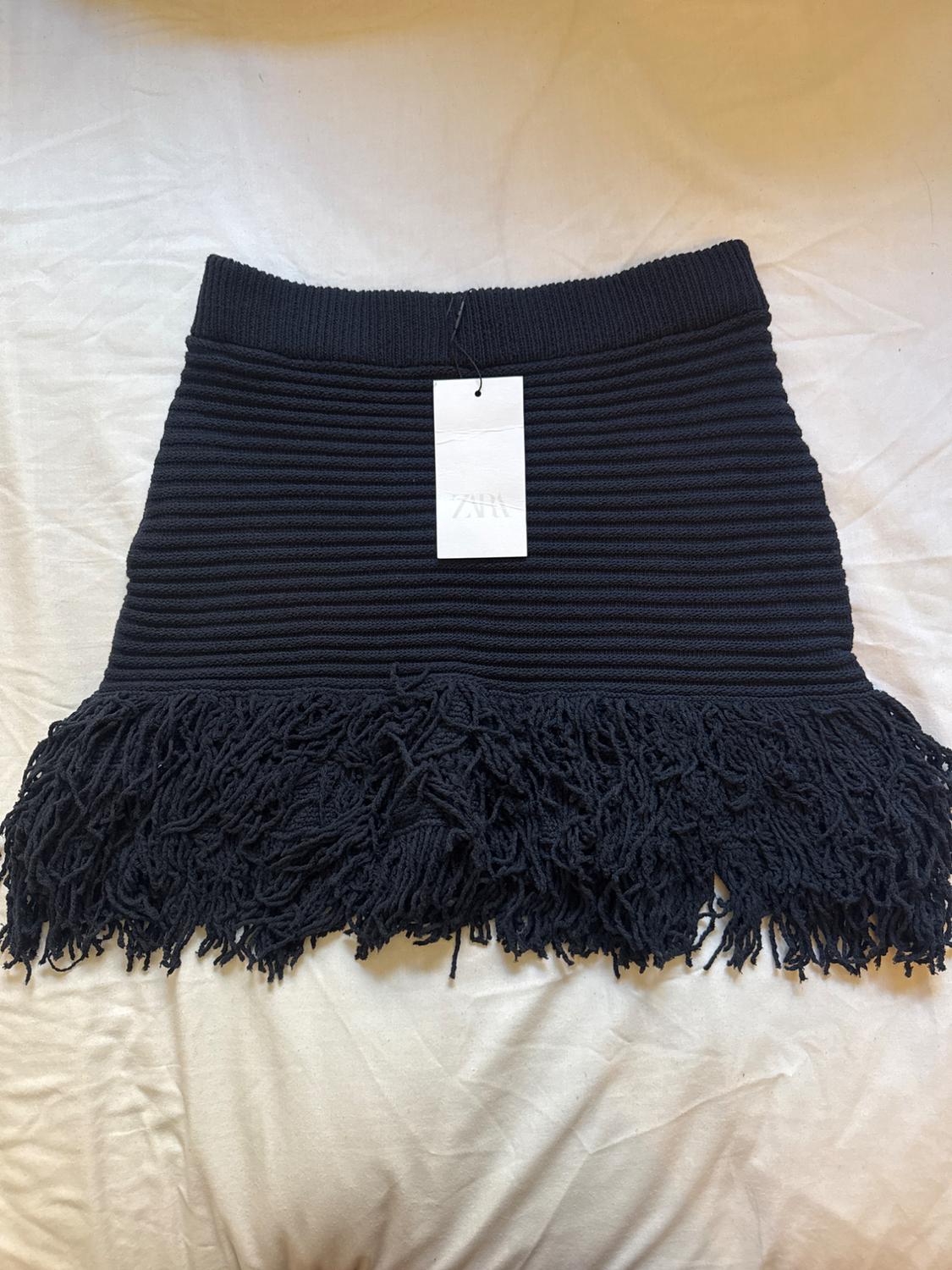Black ribbed knit miniskirt with a chunky fringe hem, laid out on a white fabric. Includes a label with the text ZARA.
