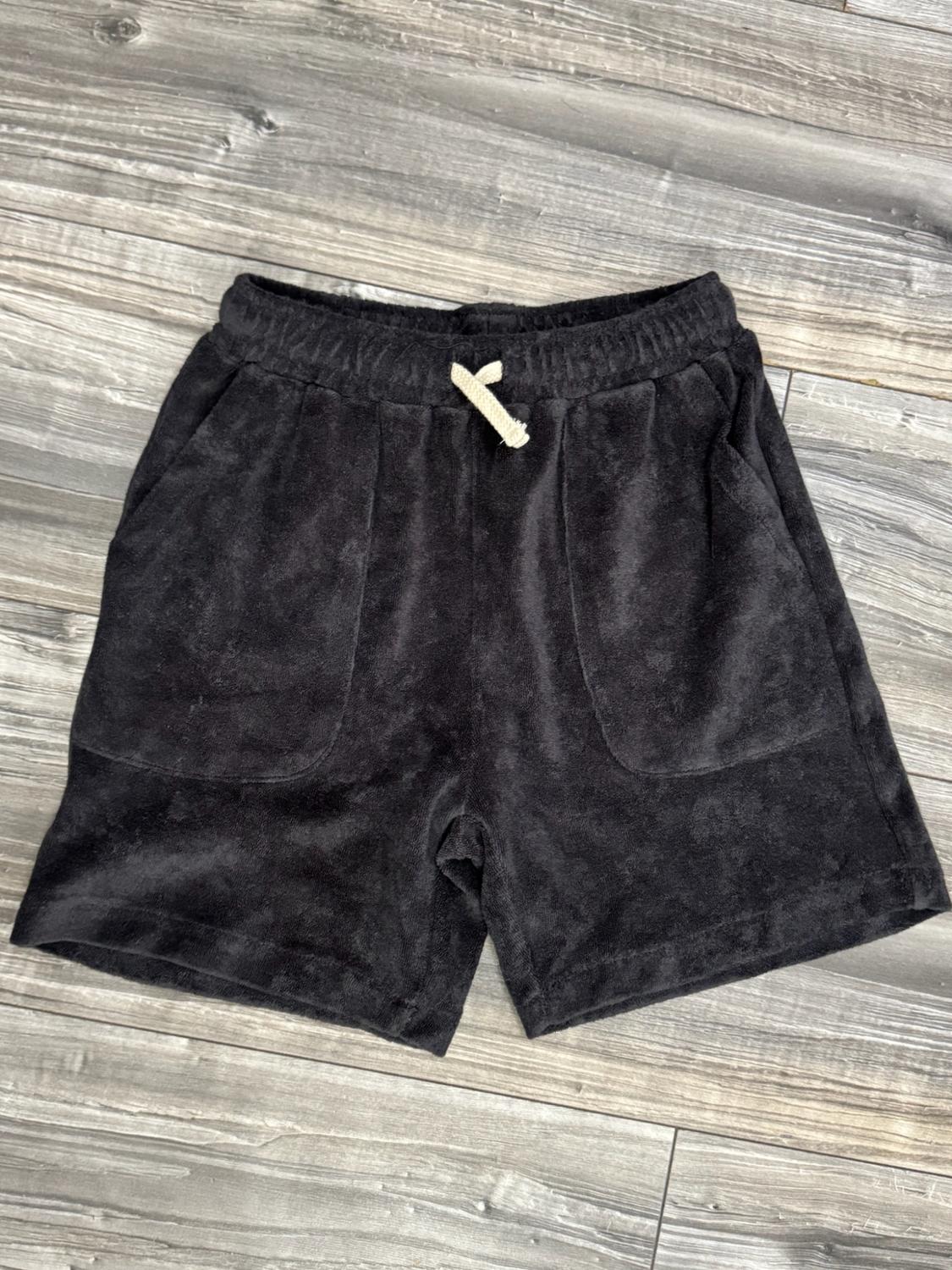 Black shorts made from soft fabric with an elasticated waistband, light-coloured drawstring and two front pockets, laid on a grey wooden floor.