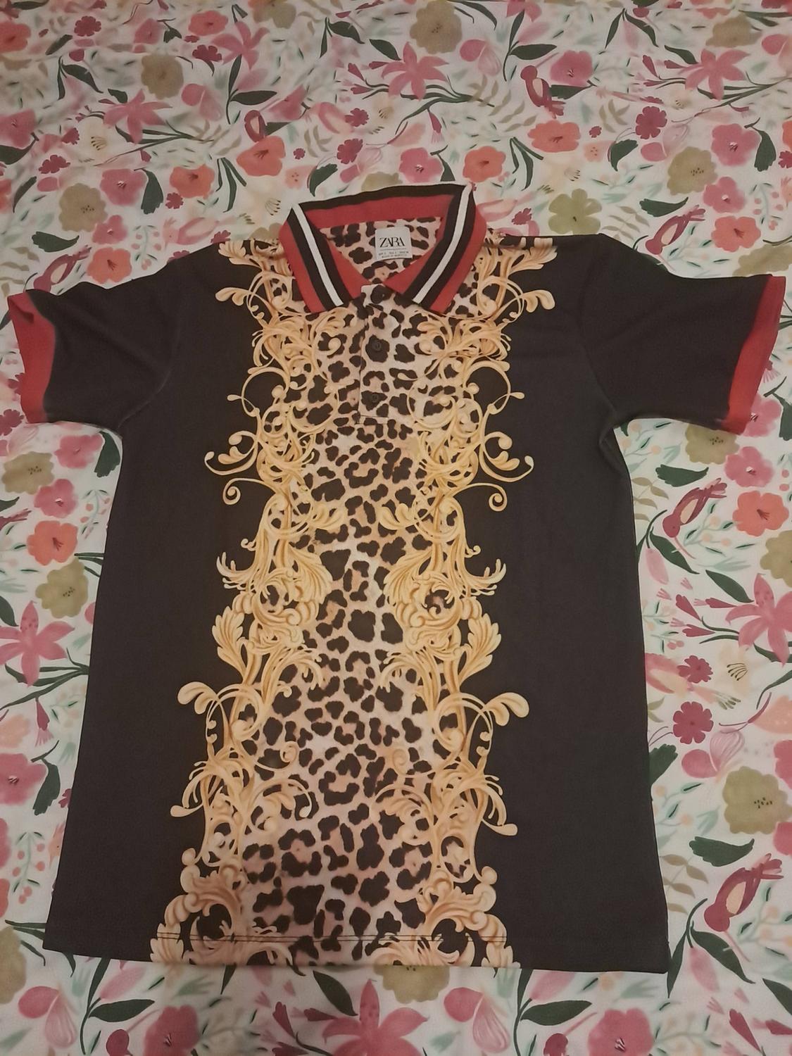 Black short-sleeved polo shirt on a floral background. It features a central leopard panel with gold details, a red striped collar and ZARA text.
