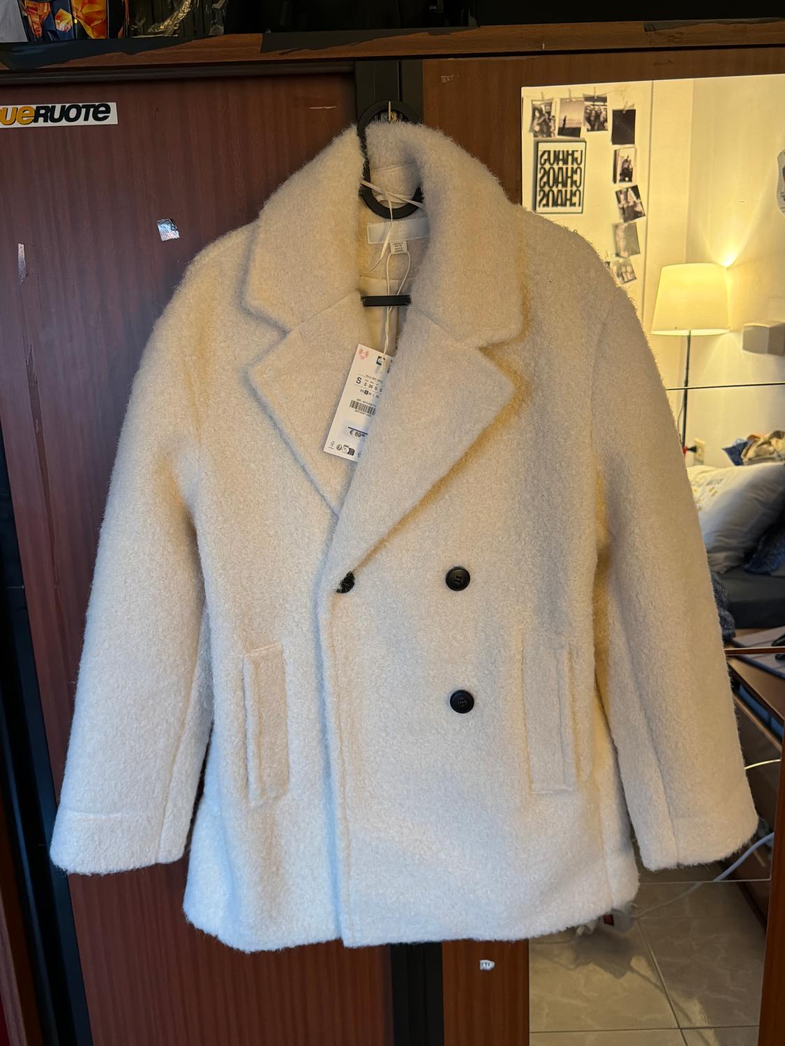 White wool coat with black buttons hanging on a hanger. Background with wooden furniture and a lit lamp.