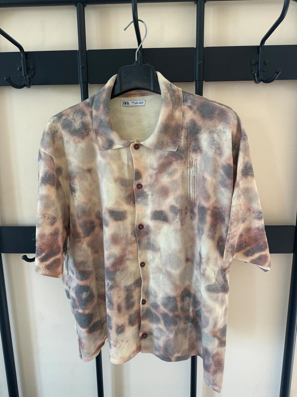 Short-sleeved shirt with an abstract print in brown and beige tones, hanging on a black hanger against a light background.