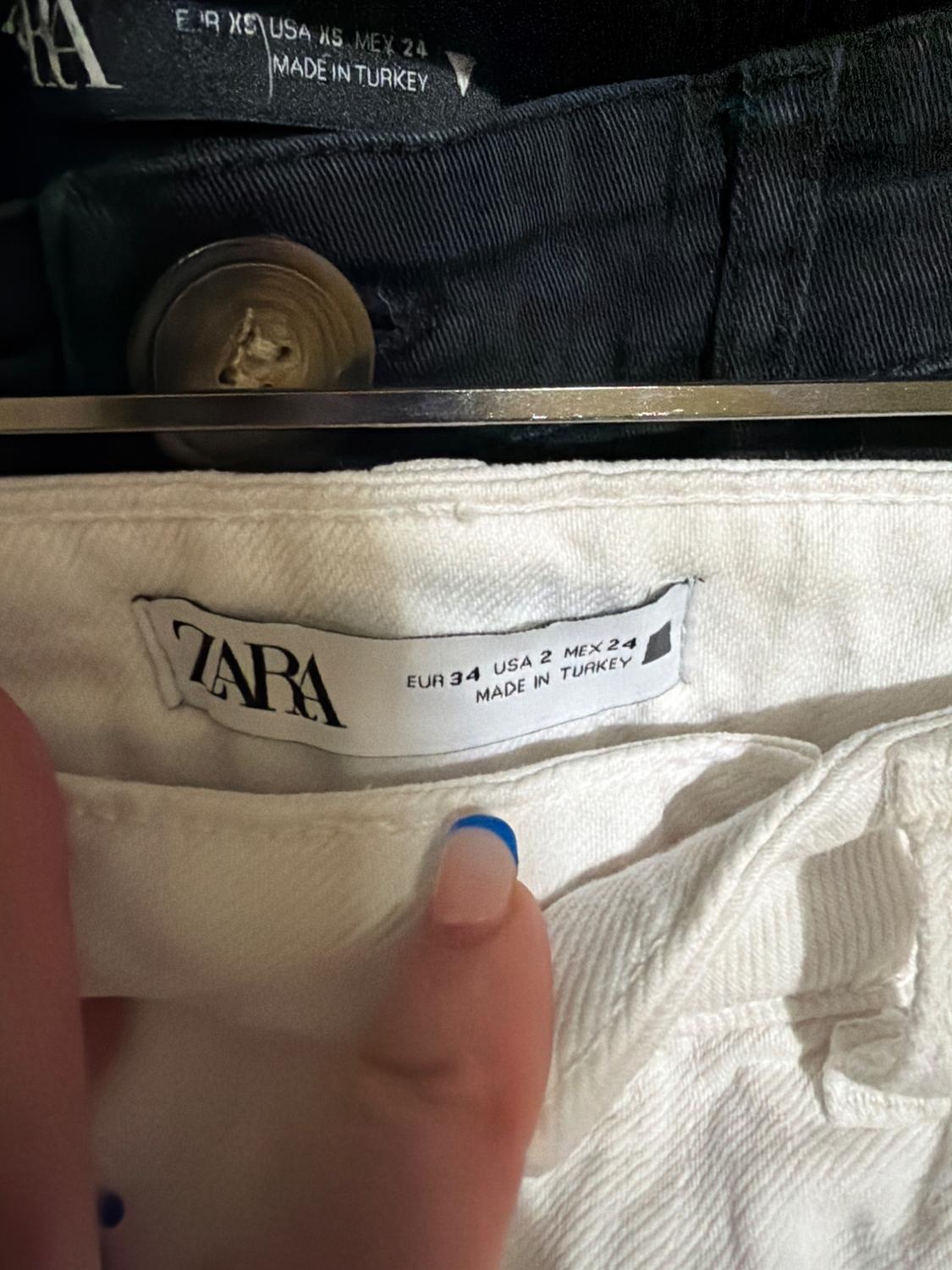 Zara white trousers label, size EUR 34, USA 2, MEX 24, made in Turkey. Background with black trousers and metallic button. Fingernail with French manicure.