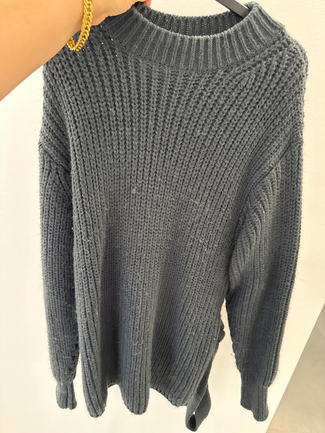 Arm with a gold bracelet holding a chunky grey knit jumper on a hanger against a white background.