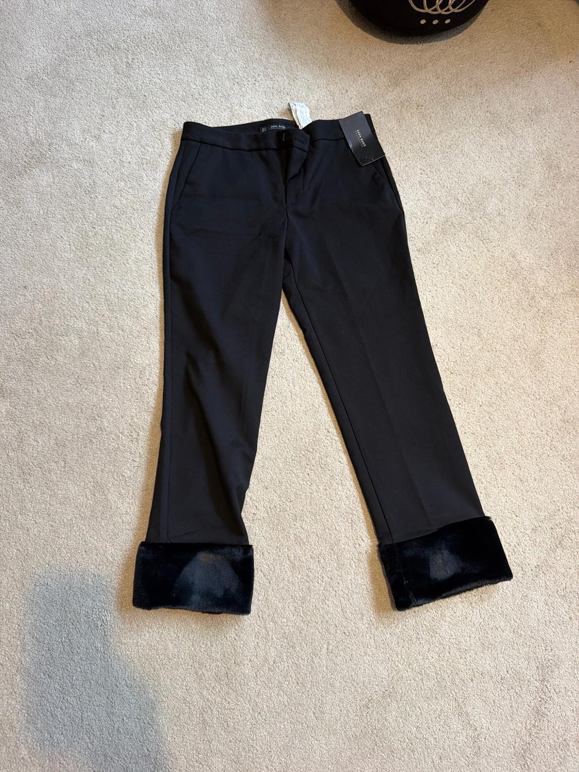 Black trousers with a black faux-fur hem, laid out on a light-coloured carpet. There are labels at the waist.