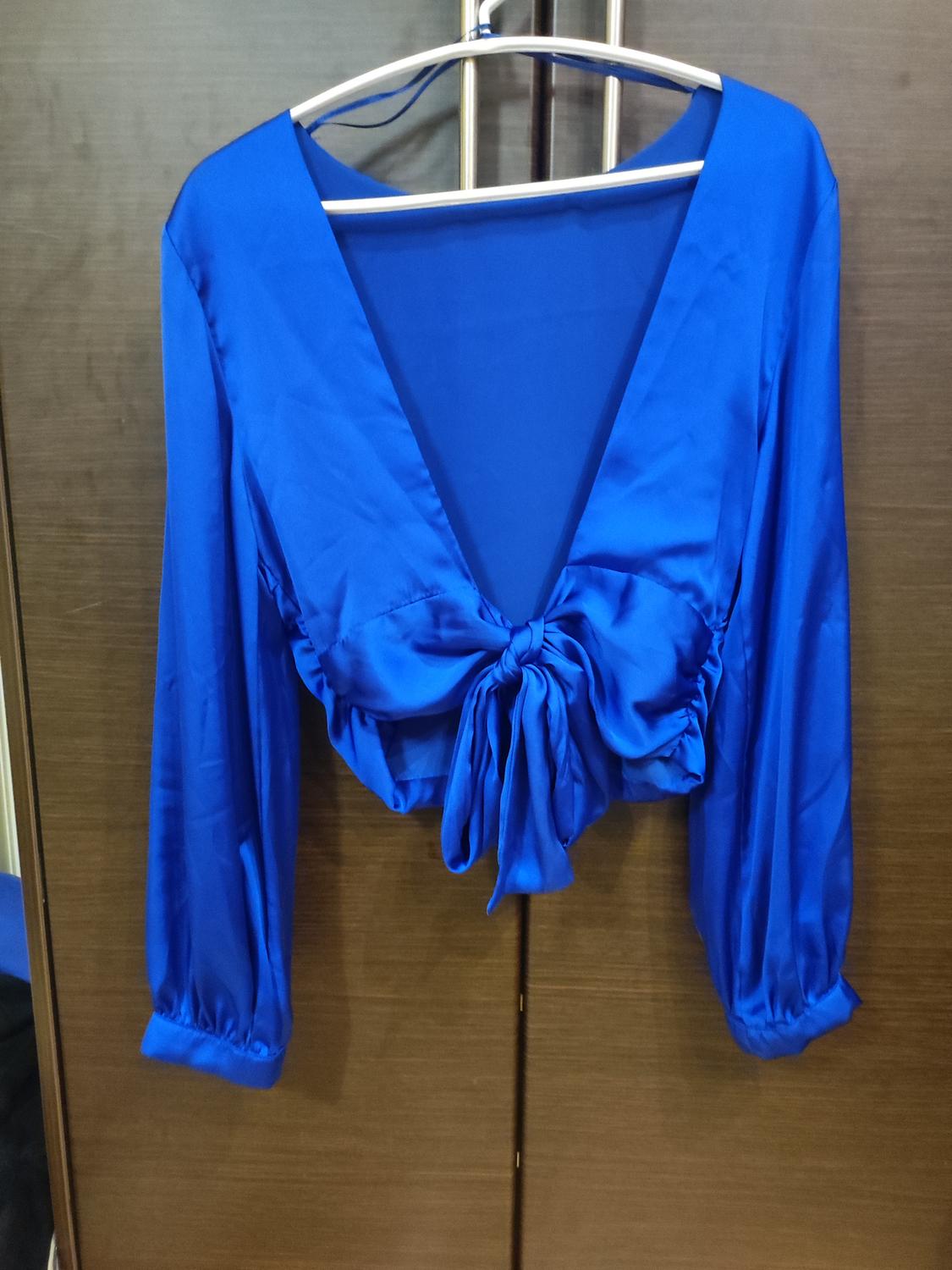 Blue long-sleeved blouse with a V-neck and front tie, hanging on a hanger against a brown wooden background.