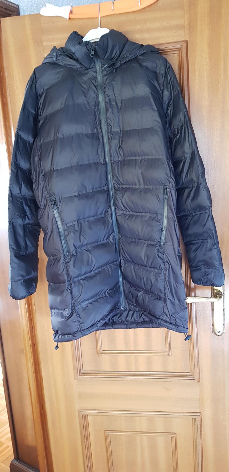 Black padded jacket with a hood, hanging on a wooden door. Visible zips on the front and side pockets.