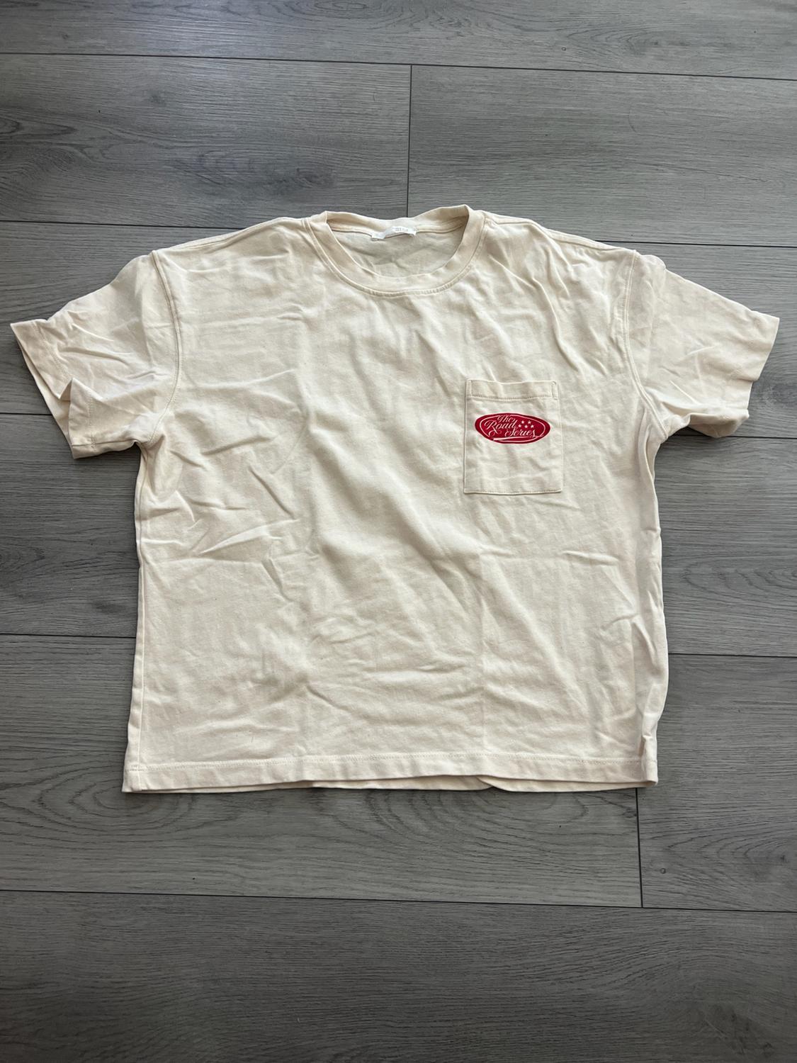 Beige short-sleeved t-shirt laid out on a gray wooden floor. Includes a pocket with a red logo and the text "The Road Series".