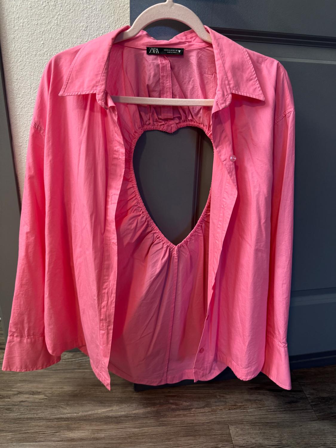 Pink long-sleeved shirt with a heart-shaped cut-out design on the chest, hanging on a hanger in front of a grey door.