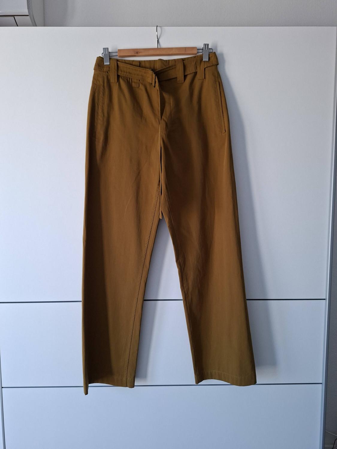 Long mustard-coloured trousers hanging from a wooden hanger with metal clips in front of a white wardrobe with horizontal lines.