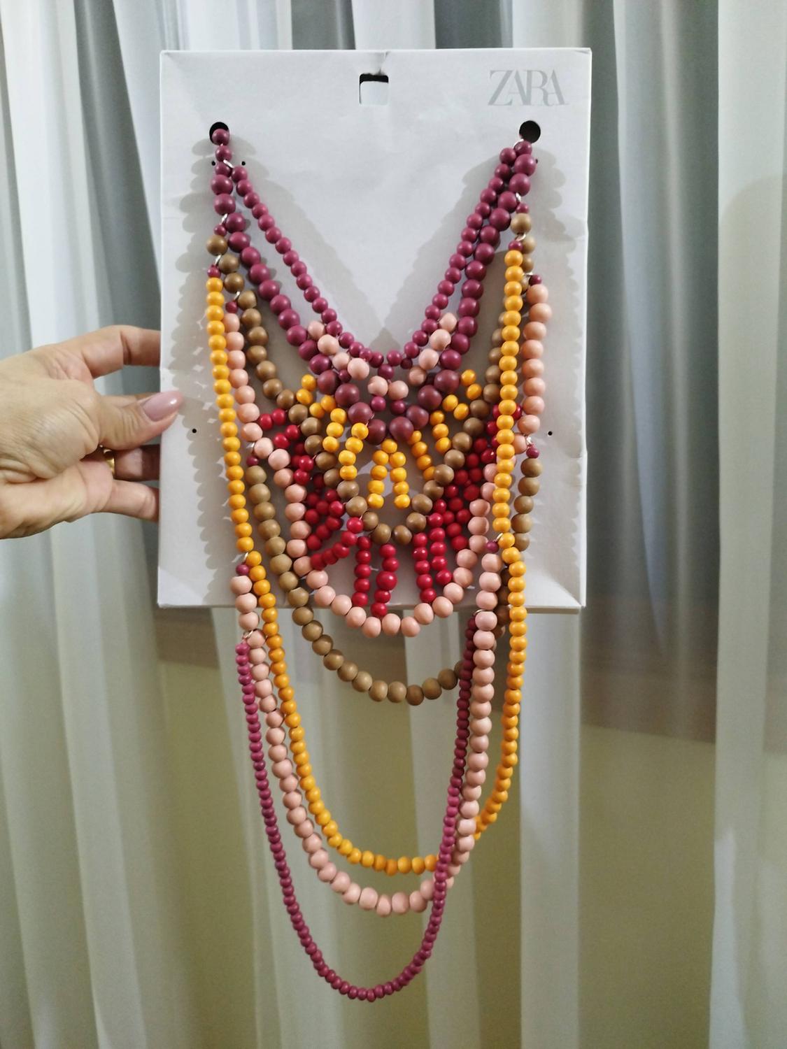 Hand holding a layered beaded necklace in burgundy, pink, orange, and brown tones against a cardboard backing with the text ZARA. Background of curtains.