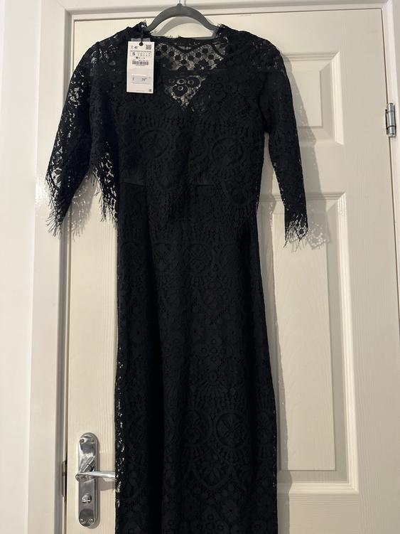Black lace dress hanging on a white door. Visible tag with price of €39. Three-quarter sleeves and floral design.