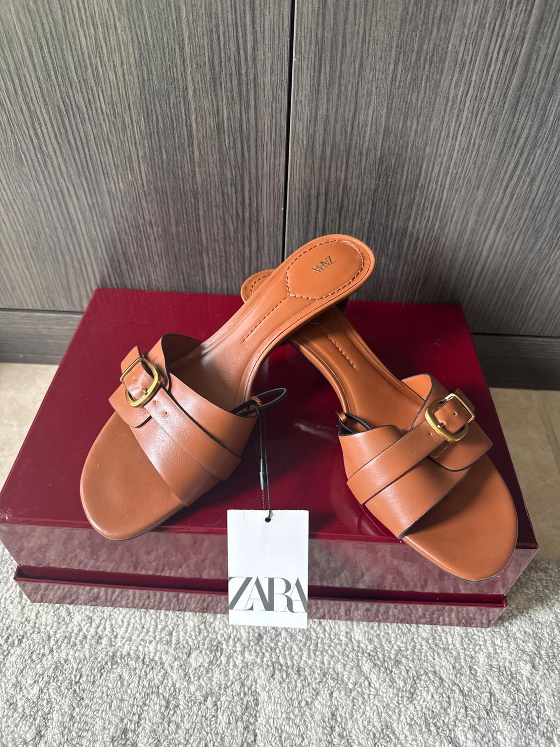 Brown flat sandals with a gold buckle on a red box. Label with the text ZARA. Wooden background and grey rug.