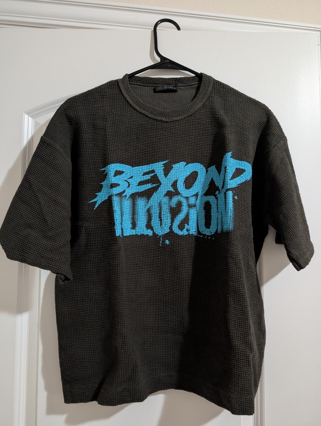 Black short-sleeved textured T-shirt, hanging on a hanger. Blue text on the front: "BEYOND ILLUSION".