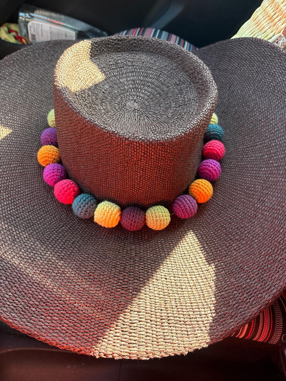 Dark brown woven hat with colourful crochet spheres on the crown, under a strong ray of sunlight casting shadows across the brim.