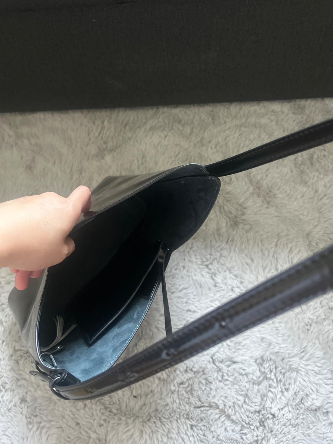 Hand opening a black bag with long handles to show its interior and pocket, resting on a soft, light-toned rug.