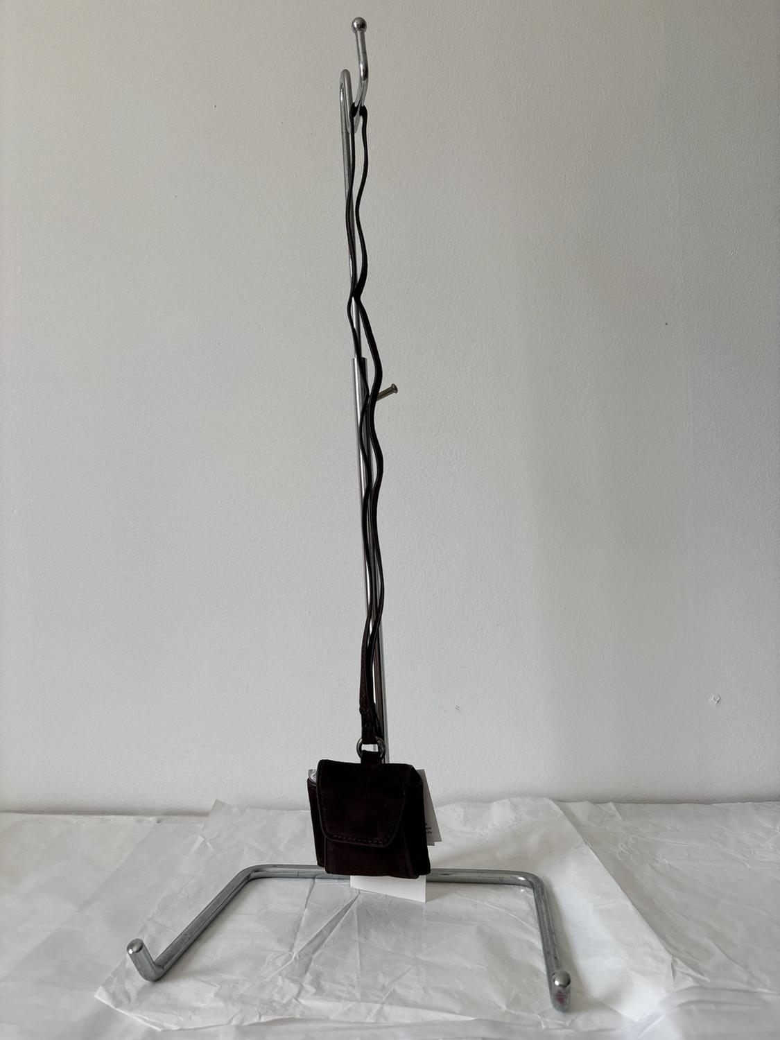 Small dark brown handbag hanging by its strap from a metal stand on crumpled white paper, in front of a white wall.