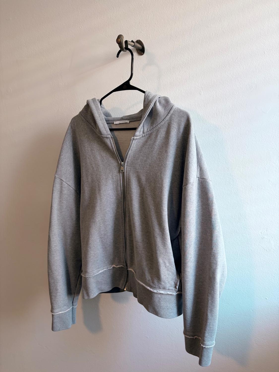 Gray zip-up hoodie with a hood, hanging on a black hanger on a wall. It has unfinished edges at the hem and cuffs.