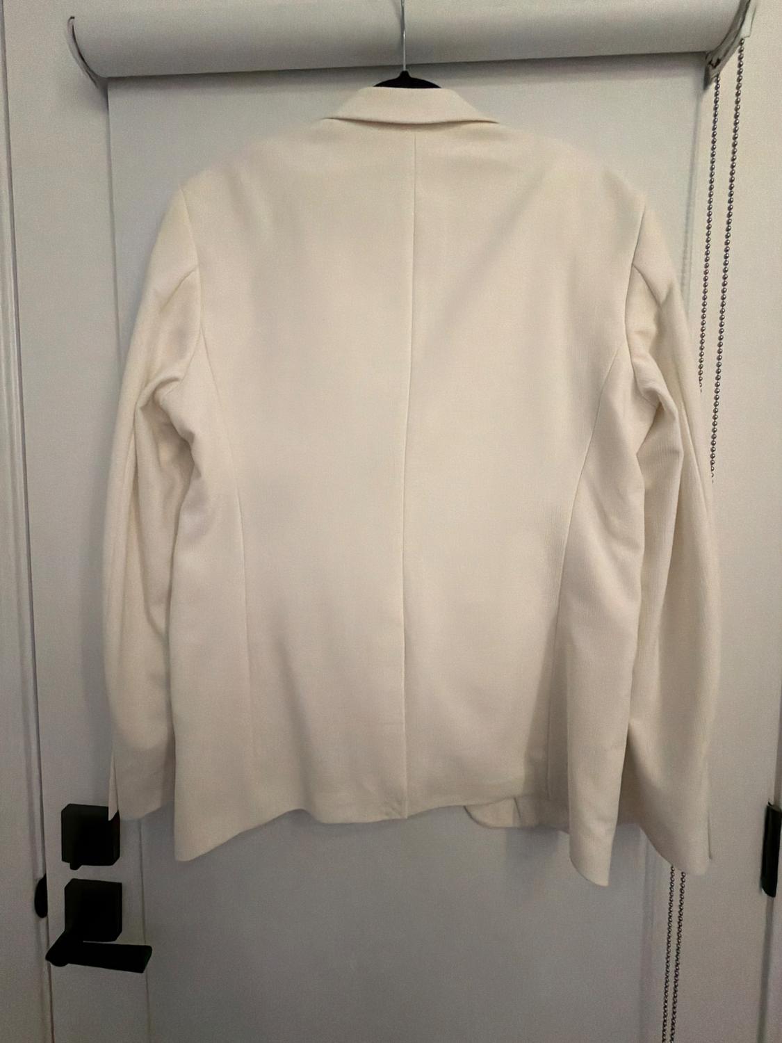 White blazer seen from behind, hanging on a black hanger on a white door indoors.