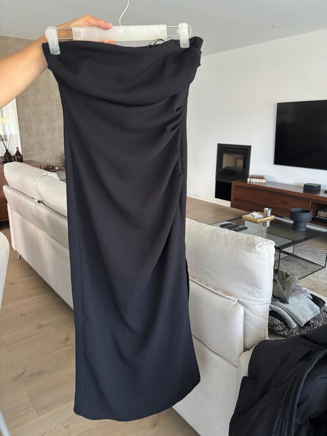 Arm holding a black gathered dress on a hanger. In the background, a modern living room with a white sofa, creating a relaxed atmosphere.