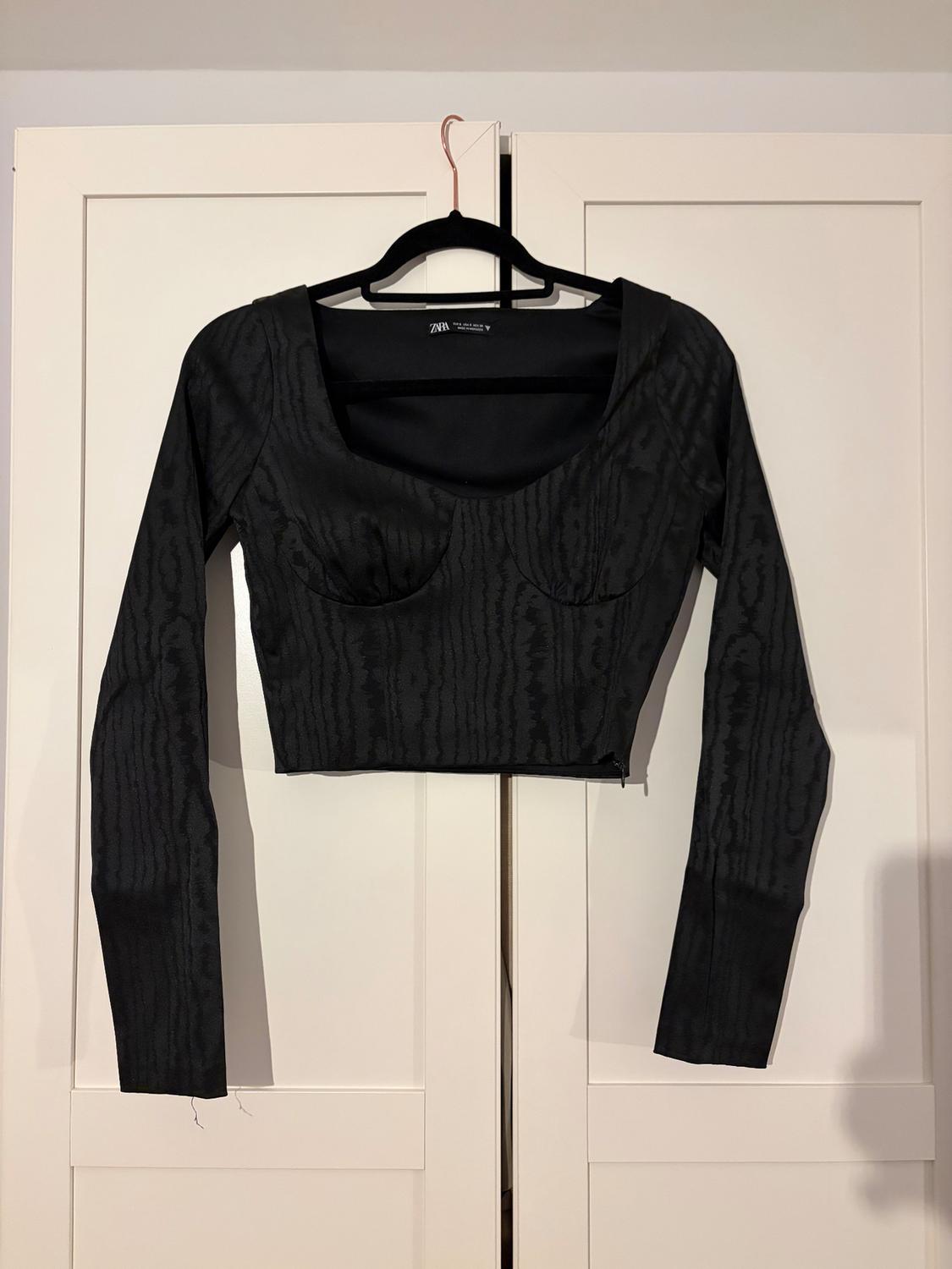 Black long-sleeved textured top, hanging on a black hanger in front of a white wardrobe. Visible text: "ZARA".