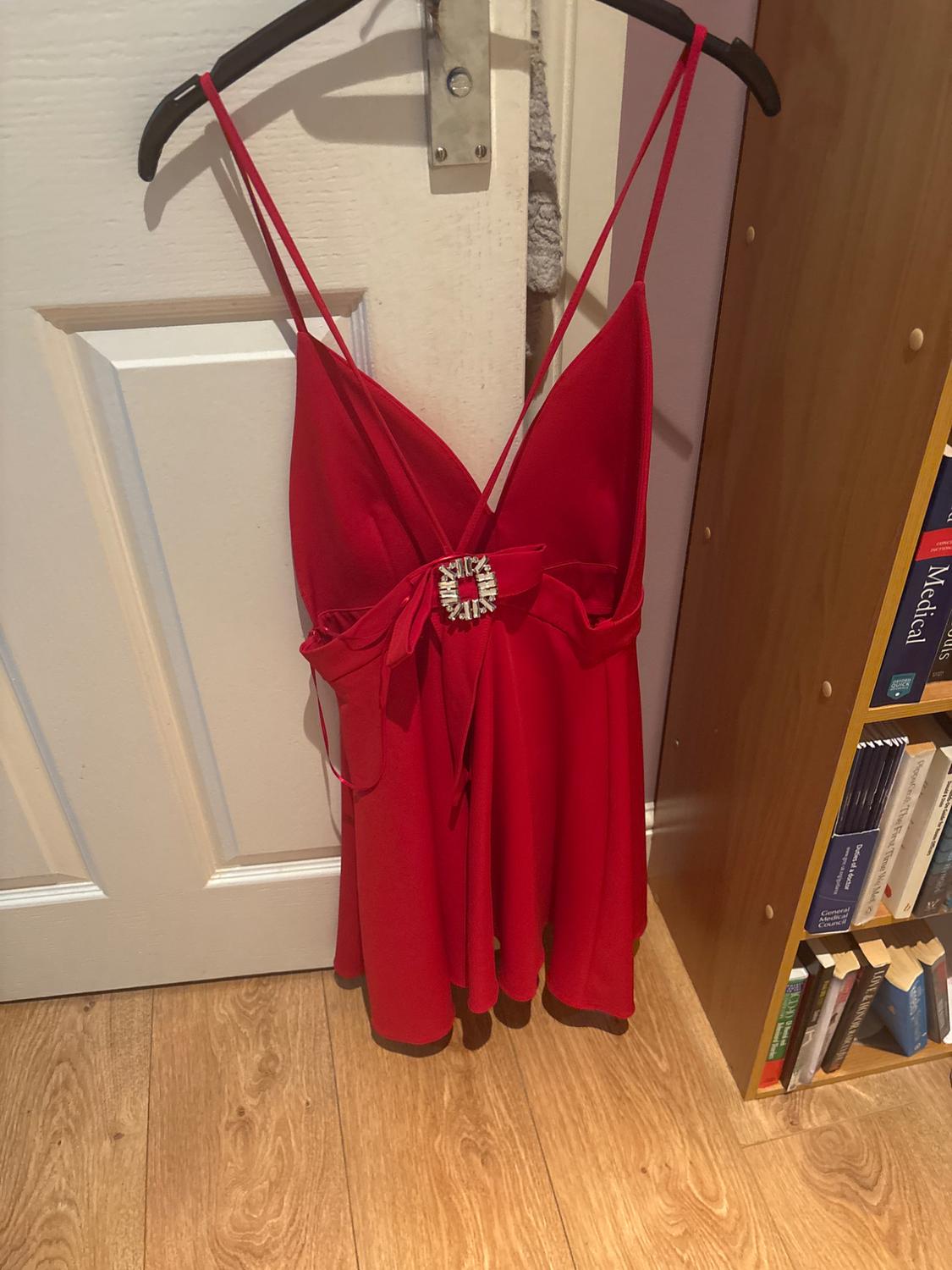 Red dress with spaghetti straps and brooch detail at the centre, hanging on a hanger in front of a white door.