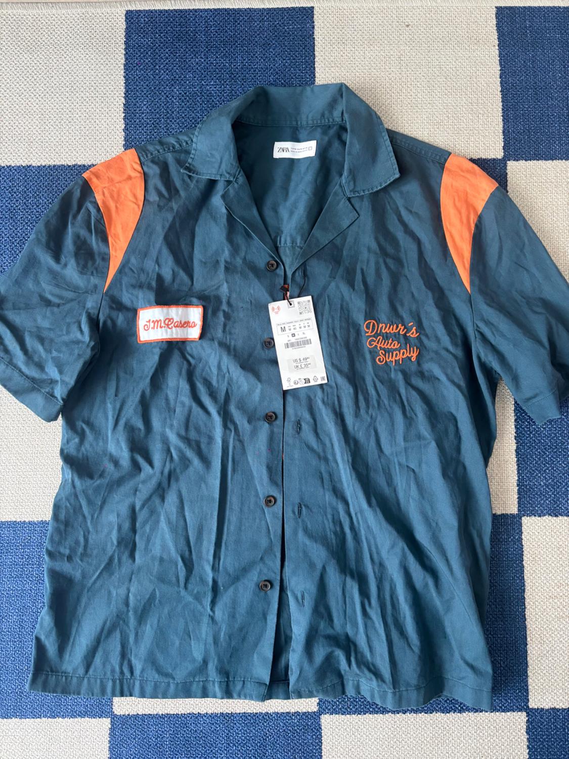 Blue shirt with short sleeves and orange accents. Visible text: "J.M. Casero" and "Dnur's Auto Supply". Hanging tag in the center.