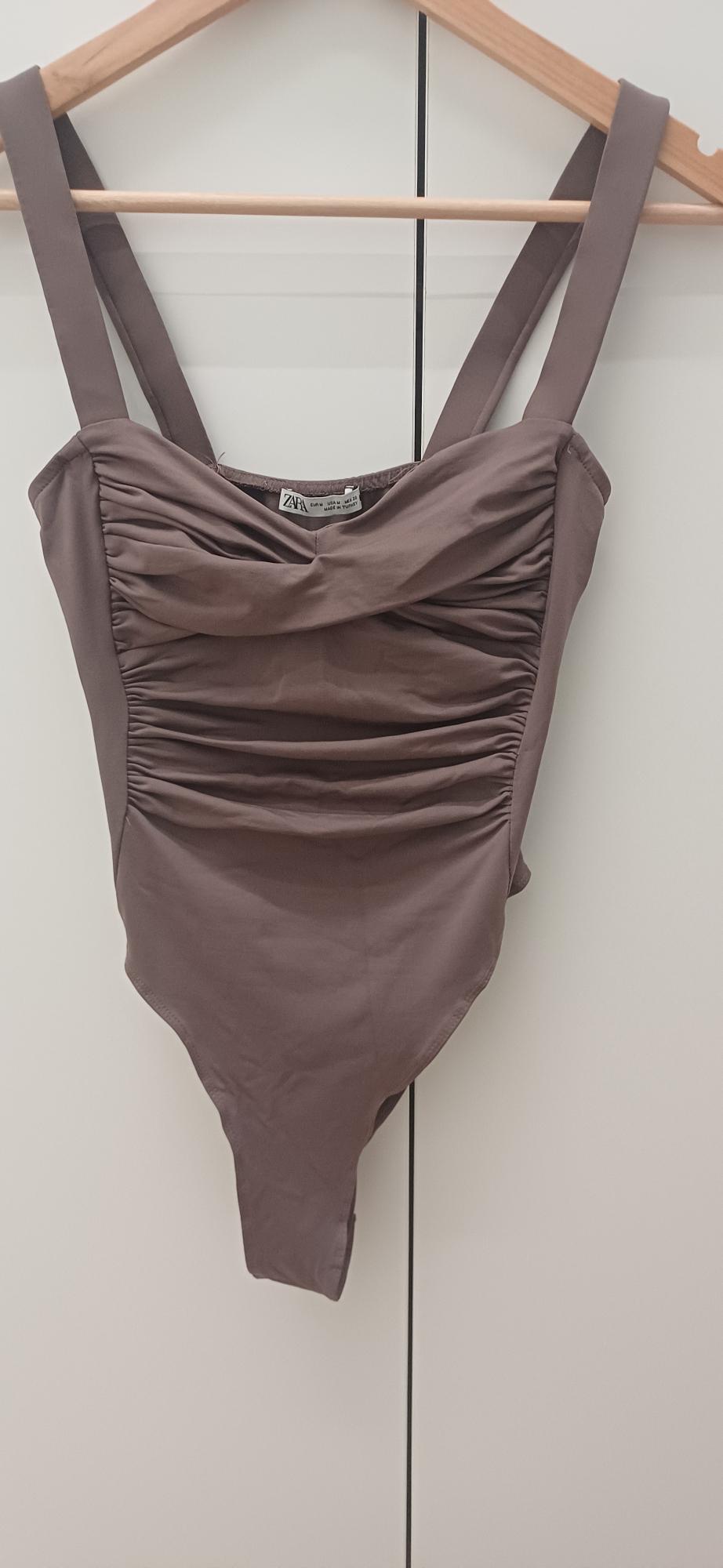 Brown bodysuit with wide straps and ruched bodice, hanging on a wooden hanger against a white background. Visible text: ZARA.
