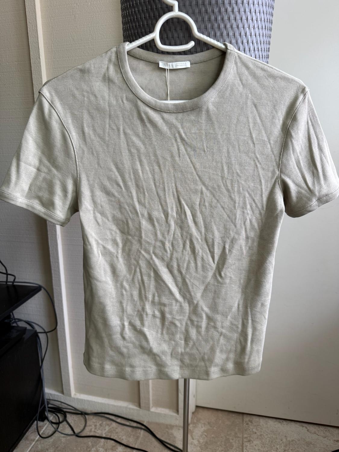Basic short-sleeved T-shirt in light beige, with visible creases, hanging on a white hanger in a domestic setting.