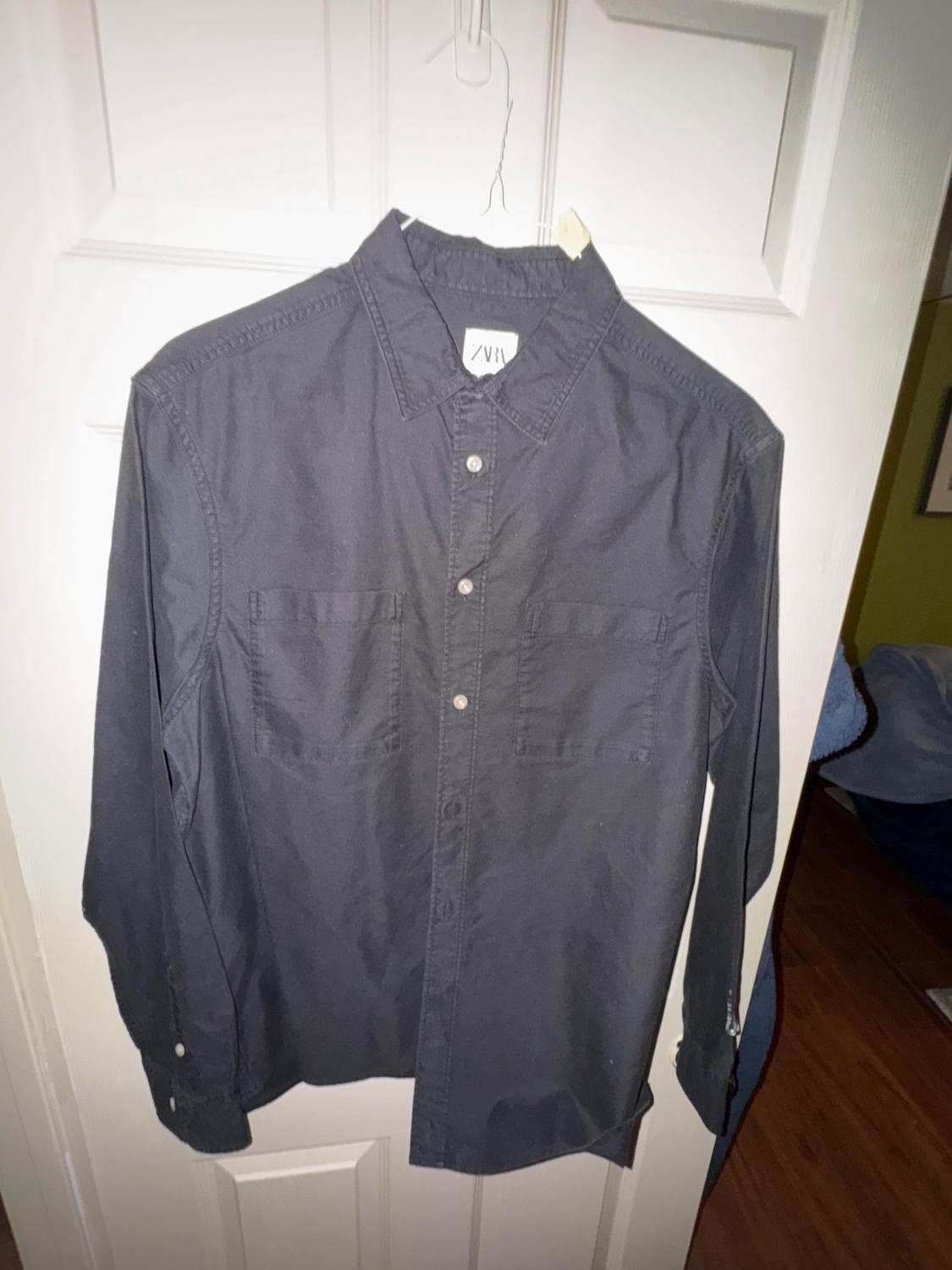 Black long-sleeved shirt hanging on a white door. It has two chest pockets and front buttons. Visible label with text "ZARA".