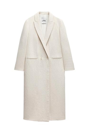 Long white textured coat with wide lapels and button closure. Visible label with the text "ZARA". White background.