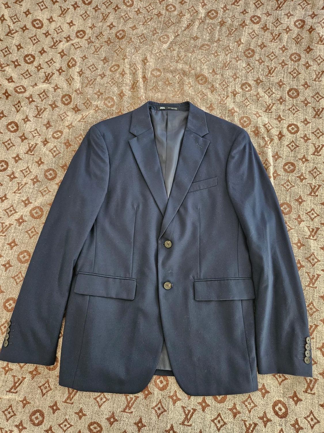 Navy blue two-button jacket with flap pockets, on a brown patterned fabric background. Text: ZARA.