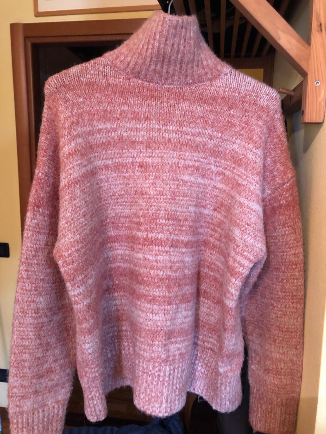Mottled pink chunky knit jumper hanging on a hanger, with a high neck, in an interior with a wooden staircase.