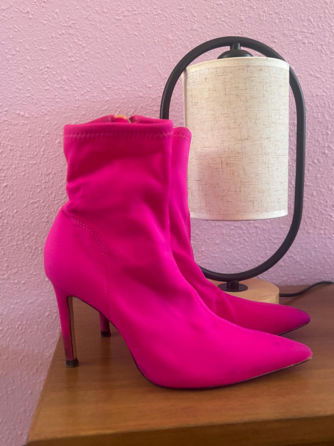 Fuchsia high-heeled boots on a wooden table, next to a lamp with a beige shade and black base, with a pink wall in the background.