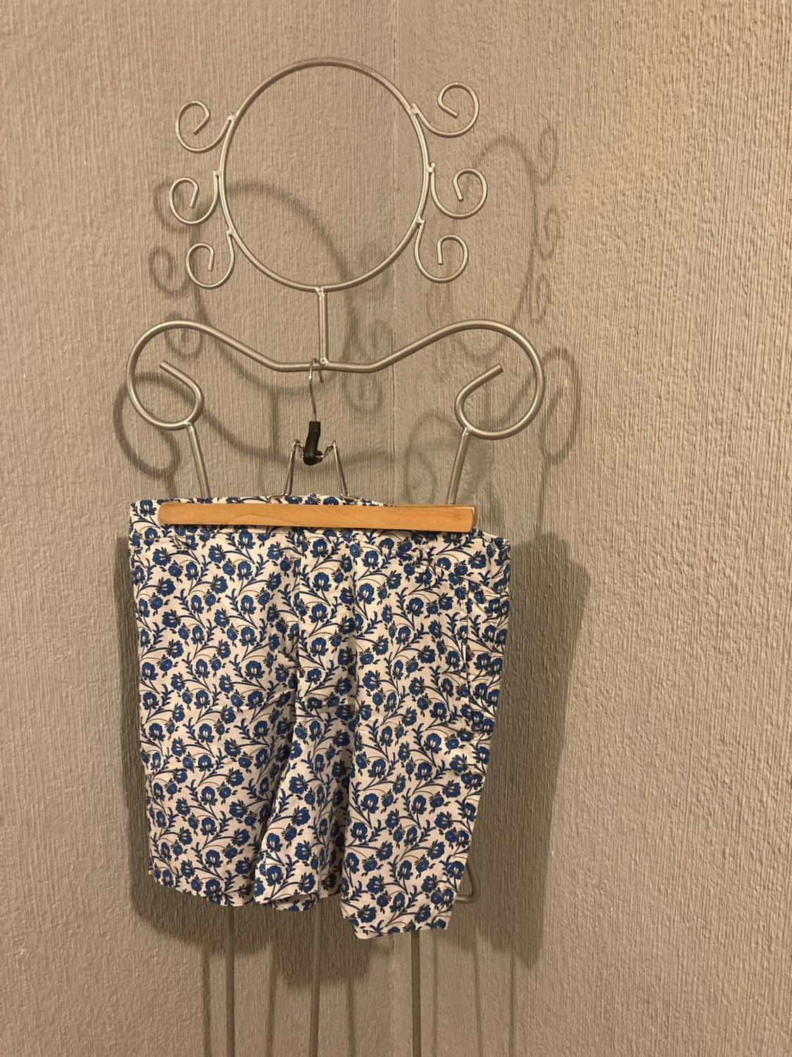White shorts with a blue floral print, hanging on a decorative wire hanger against a textured grey wall.