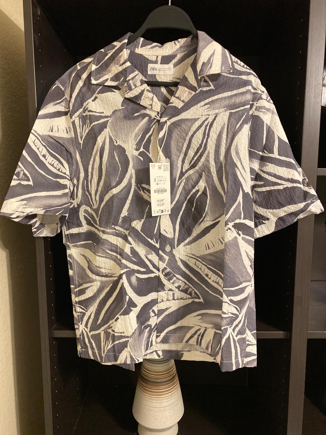 Short-sleeved shirt with a leaf print in grey and white tones, hanging on a black hanger with a visible tag.