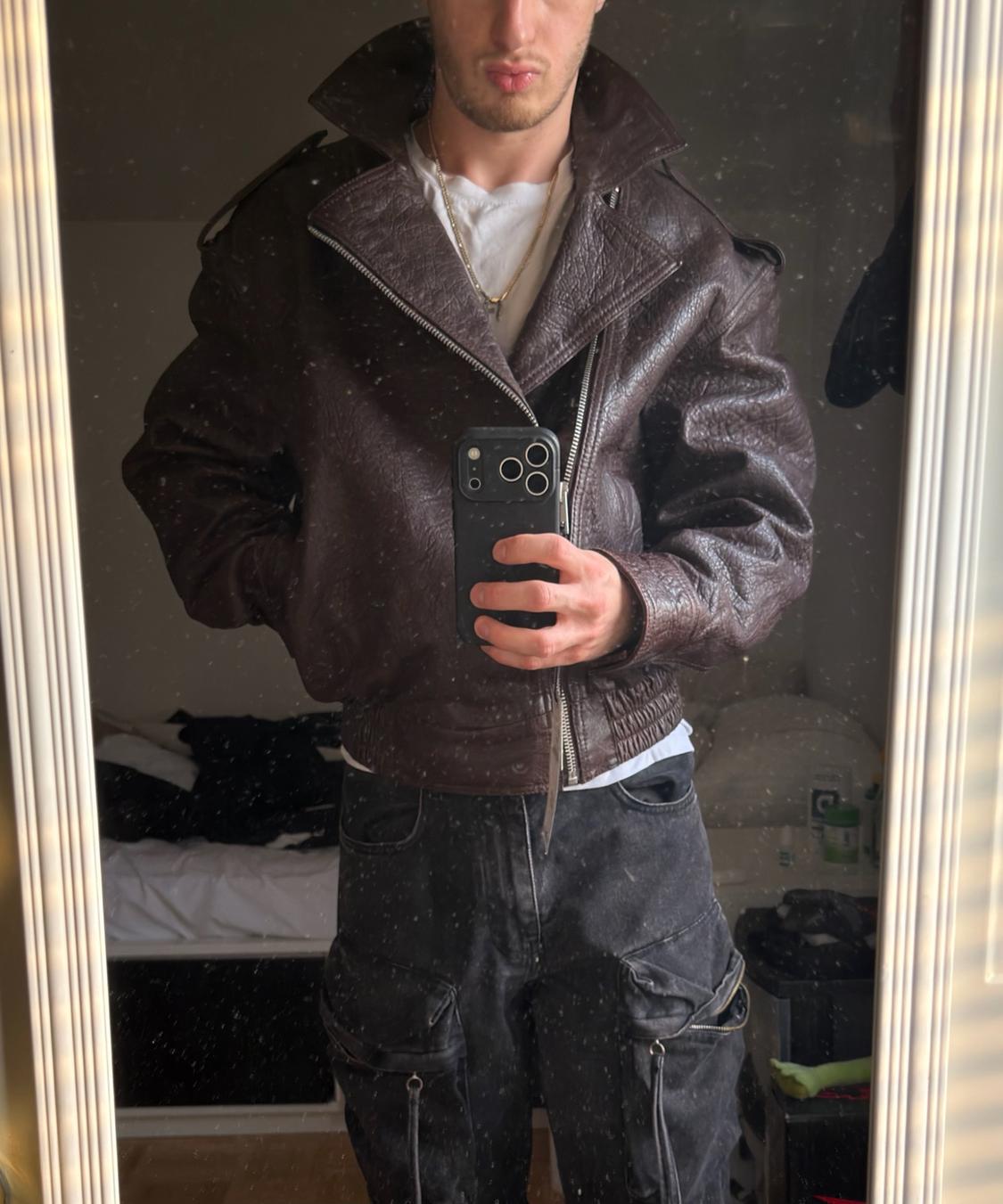 Mirror selfie in a room. Man wearing a textured brown jacket, gold necklace, white t-shirt, and black cargo pants.