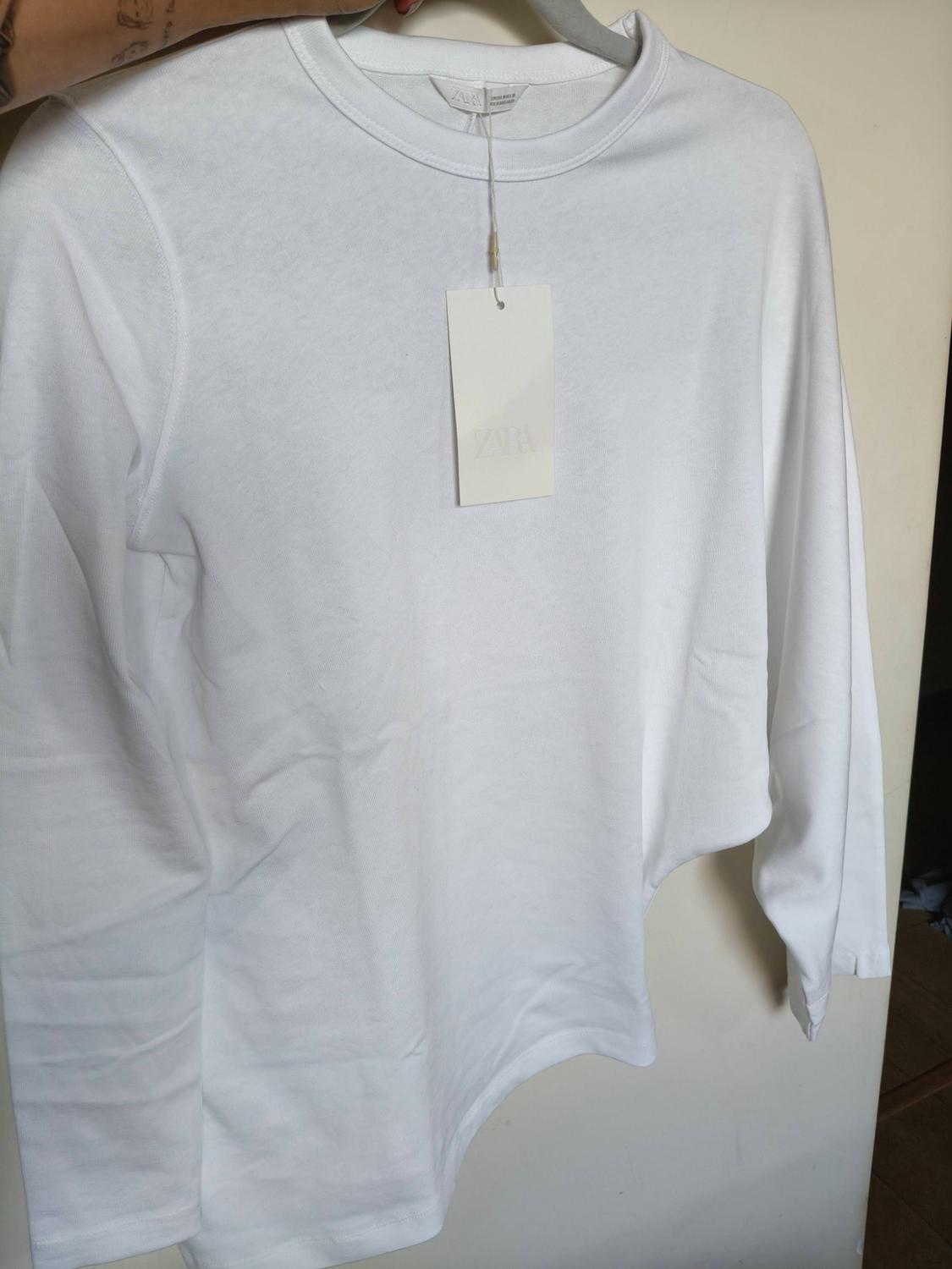 A white long-sleeved T-shirt hangs from a hanger held by a tattooed hand. A label bearing the inscription ZARA stands out.