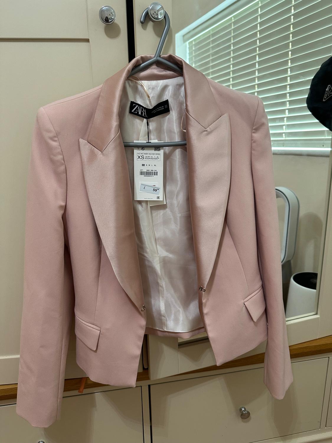 Light pink blazer hanging on a hanger with a price tag of €59.95. Beige wardrobe background with a white blind reflected.
