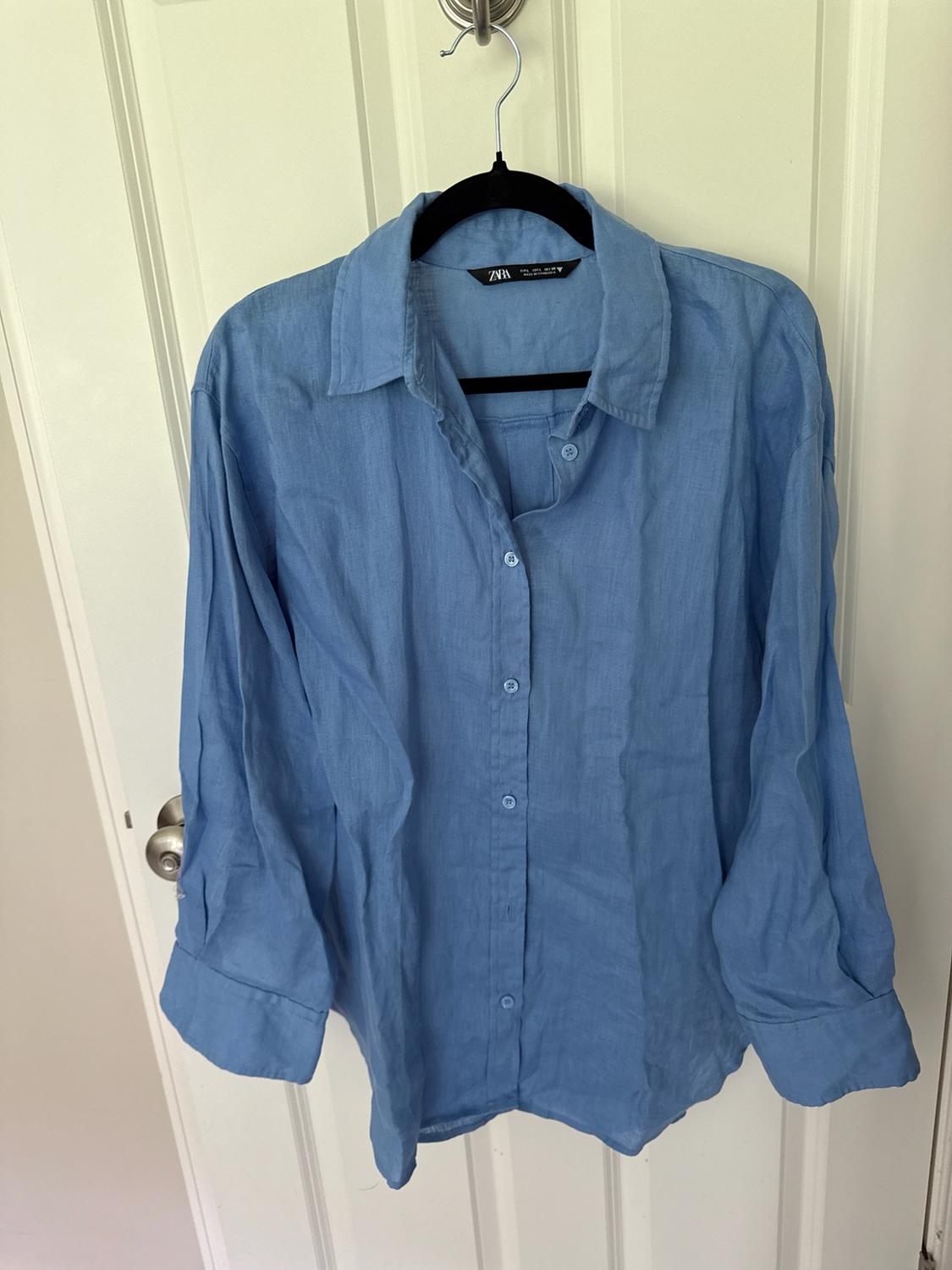 Long-sleeve blue button-up shirt hanging on a black hanger over a white door. Tag with ZARA text.