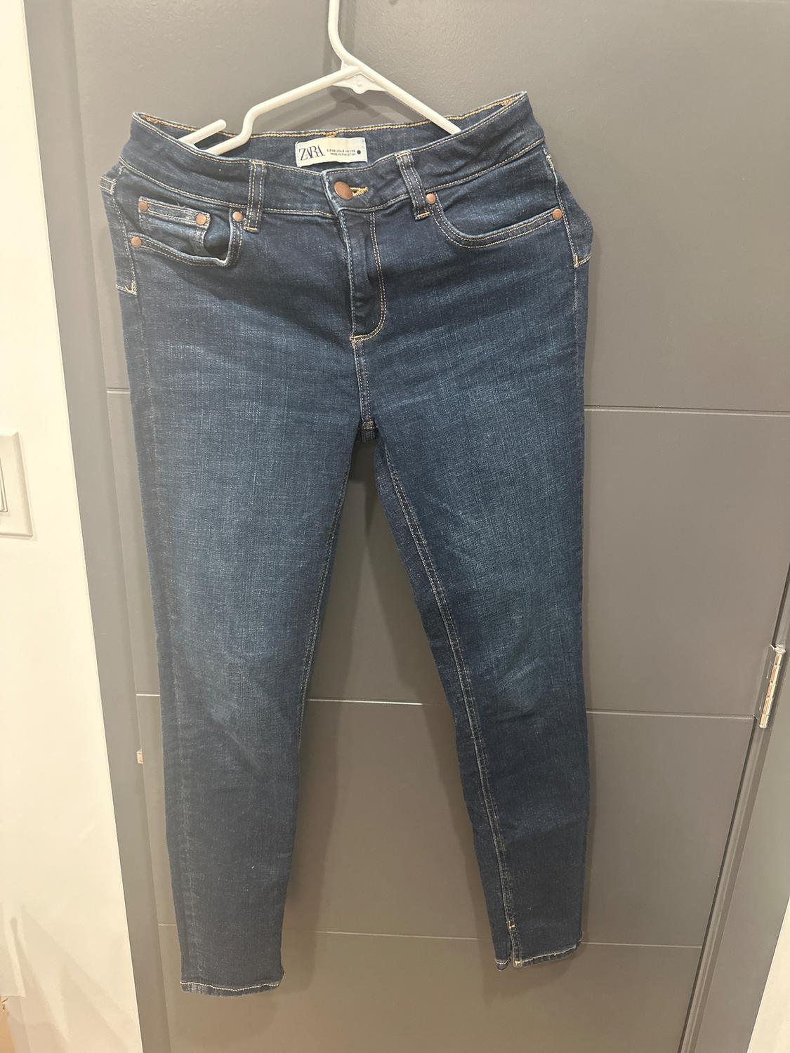 Dark blue jeans hanging on a white hanger over a gray door. Visible label: "ZARA".