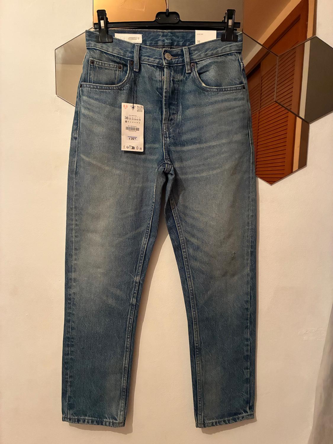 Light blue jeans hanging on a black hanger, with a visible label showing size 36, against a white background and hexagonal mirror.