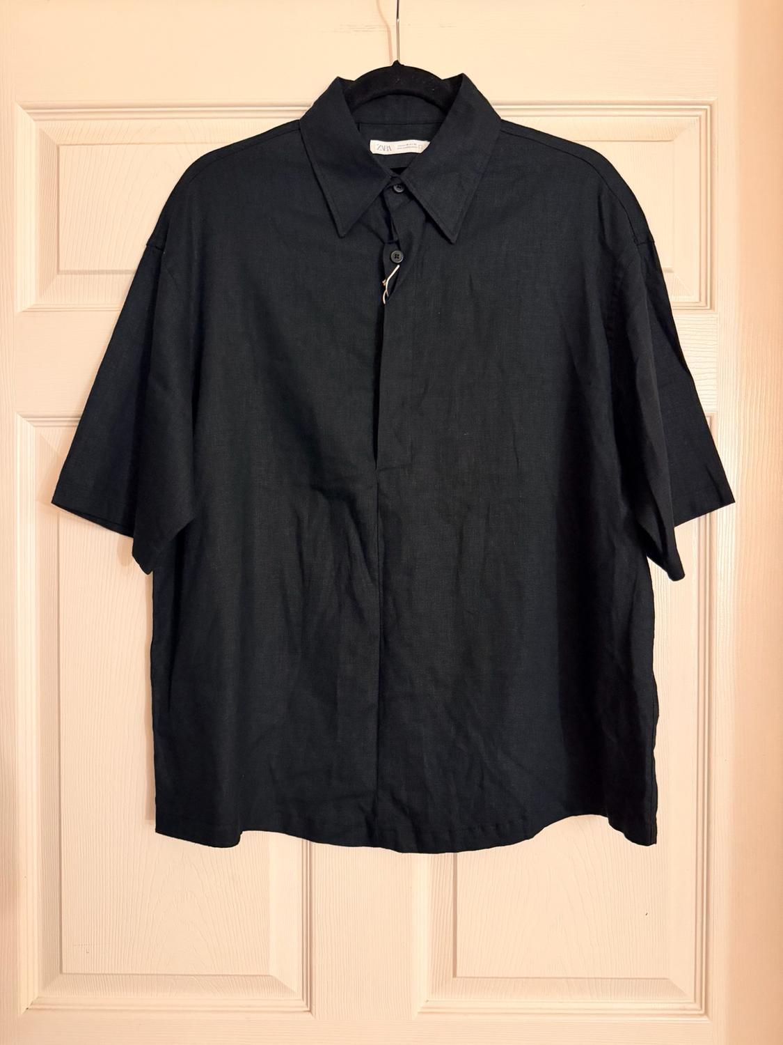 Short-sleeve black shirt hanging on a black hanger in front of a light wood door. Text: ZARA.