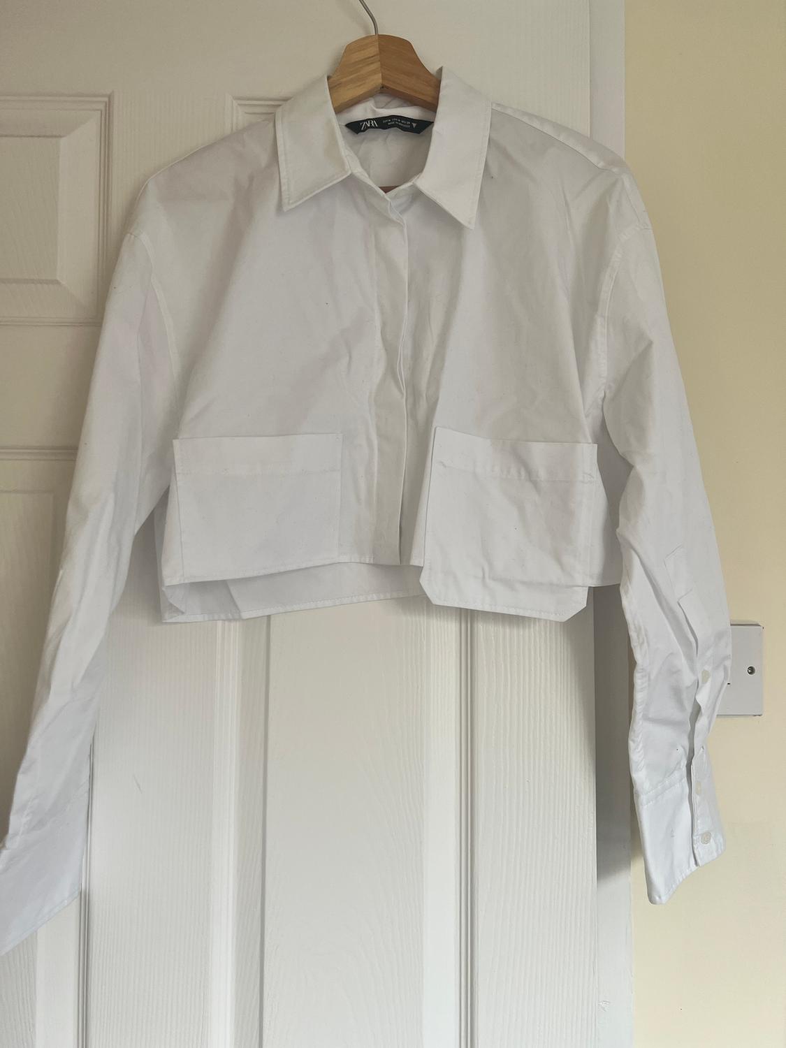 White cropped shirt hanging on a wooden hanger over a white door. It has front pockets and a classic collar.