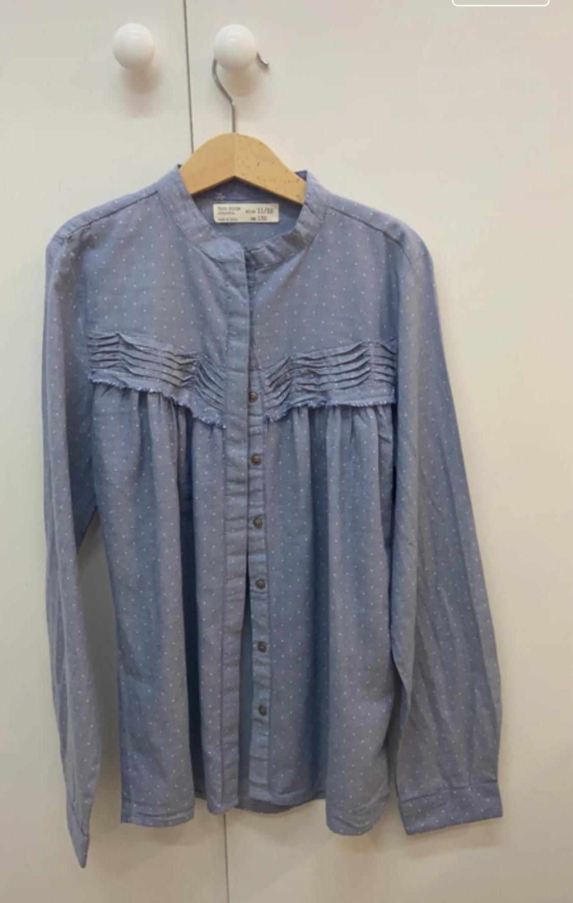 Front view of a blue shirt with white polka dots, a round neck and pleats on the chest.