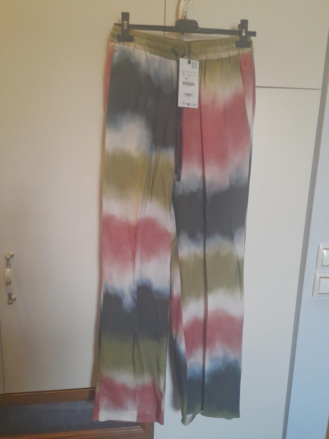 Trousers hung with a tie-dye print in red, green, and blue tones. Visible label with product details. Light wardrobe background.