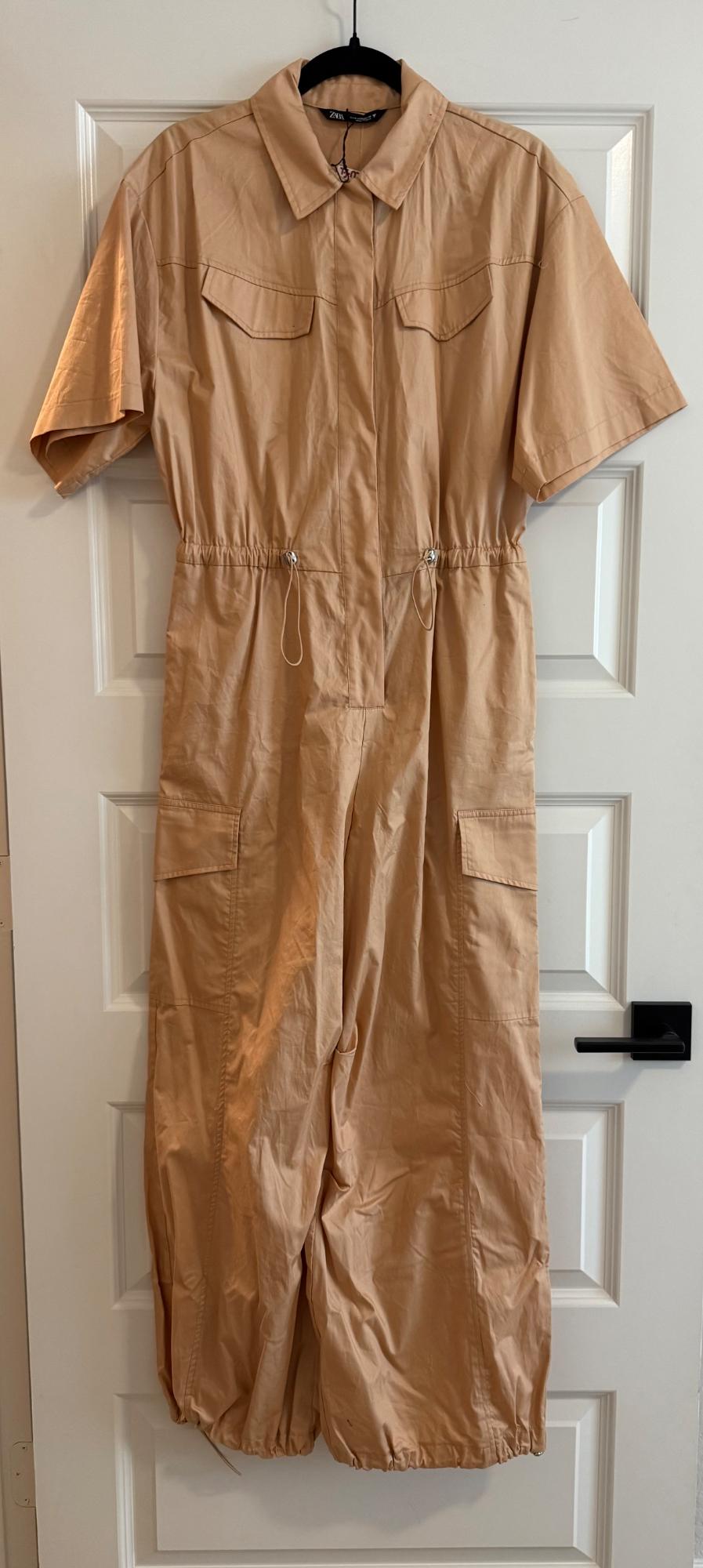 ZARA short-sleeved beige jumpsuit, with a shirt collar, cargo pockets, and an adjustable waist, hanging on a white door. Relaxed style.