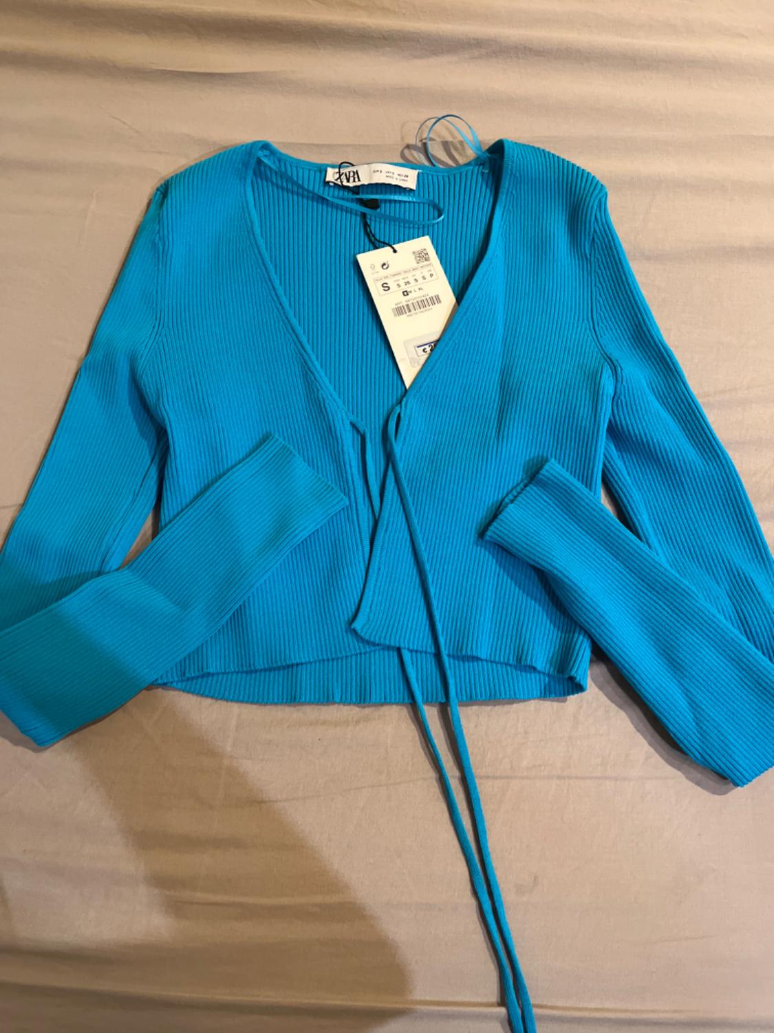 Blue ribbed knit cardigan with hanging tag on a beige surface. Long sleeves and tie at the front.